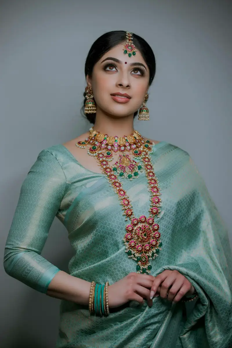 Woman in a green silk saree wearing a long gold temple necklace and jhumkas